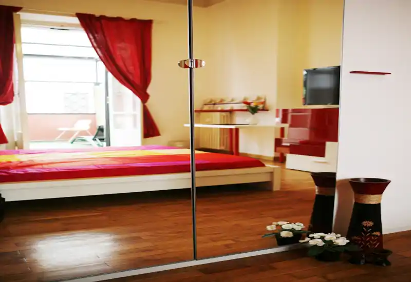 Bright, colorful bedroom with a low platform bed and large mirrored wardrobe that visually expands the space. Red curtains and warm wooden flooring create a cozy, inviting atmosphere with natural light from the balcony.
