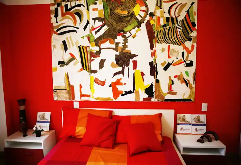 Bright, stylish bedroom with a colorful large painting above the bed and warm red-orange bedding. Two bedside tables and decorative accents create a cozy, well-styled space ideal for rental photos.