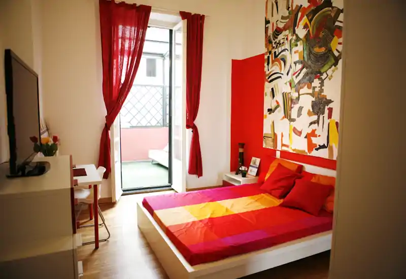 Bright, stylish bedroom with a colorful double bed, bold artwork above the headboard and a small balcony letting in natural light — ideal for a cozy city stay.