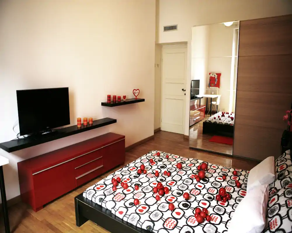 Camera da letto luminosa e moderna con copriletto decorato, tocchi rossi d’arredo e un'ampia armadio con specchio che aumenta profondità e luce.