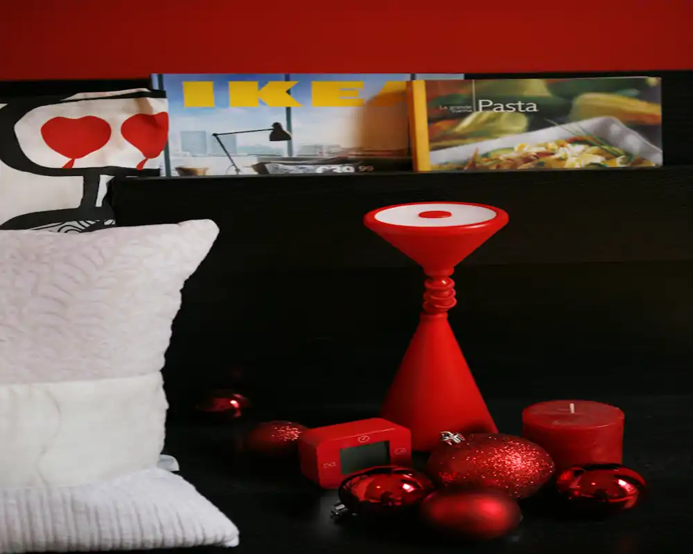 Primo piano del comodino di una camera da letto con cuscino, testiera e accenti decorativi rossi. La scena mette in risalto tessili accoglienti e oggetti d'arredo moderni in tonalità calde.