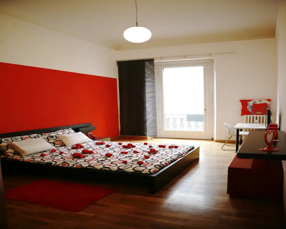 Ampia camera da letto moderna con letto matrimoniale basso su piattaforma, parete accentuata rossa e ampia porta sul balcone che illumina la stanza.