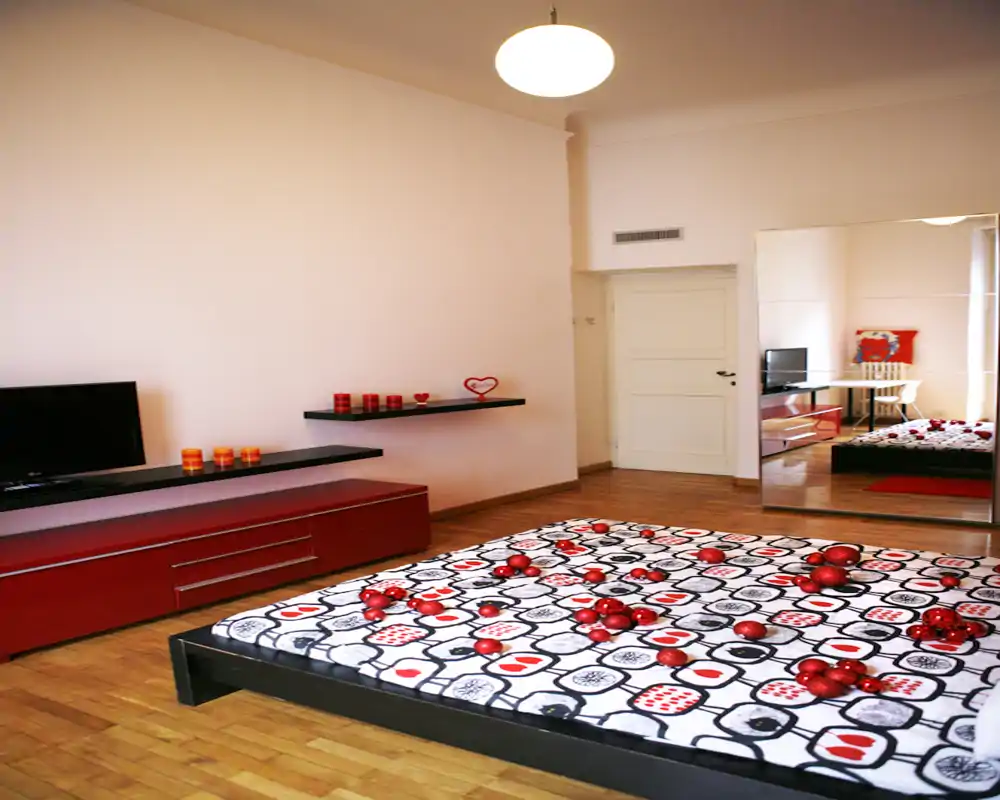 Ampia e raffinata camera da letto con letto basso su piattaforma e copriletto a fantasia, pavimento in legno caldo e dettagli rossi coordinati. Un grande specchio amplifica lo spazio e la stanza è ben illuminata con mensole moderne e mobile TV.