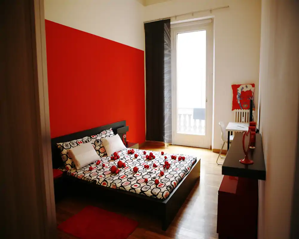 Camera da letto luminosa e di design con un letto basso matrimoniale davanti a una parete rossa, biancheria a fantasia e luce naturale dalla porta balcone.