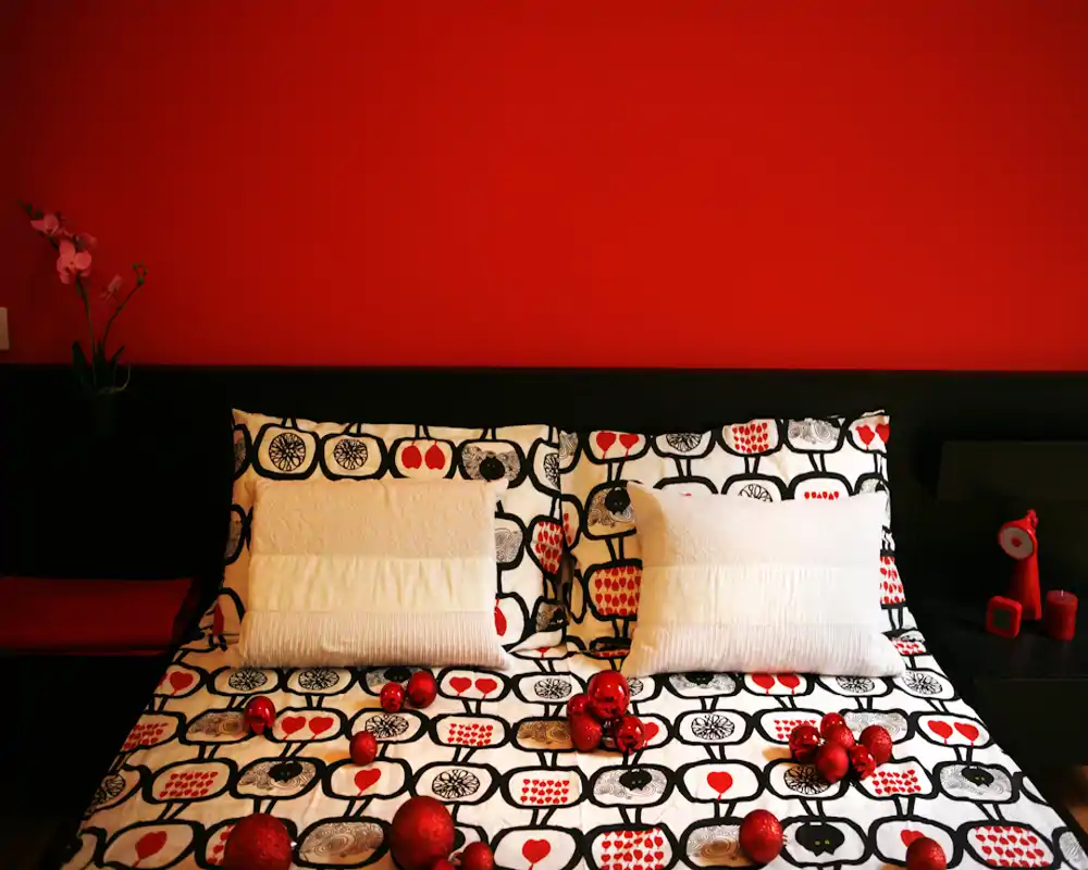 Camera da letto luminosa e di design con copriletto a motivo, due cuscini e accenti decorativi rossi su una parete rossa intensa — accogliente e ben arredata per affitti.