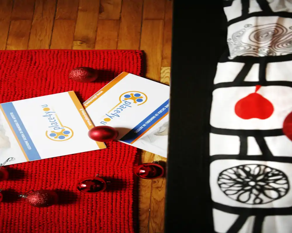 Primo piano di un tappeto rosso in maglia su pavimento in legno con palline decorative e volantini; sul lato destro si intravede il bordo di una coperta decorata, suggerendo un angolo camera da letto.