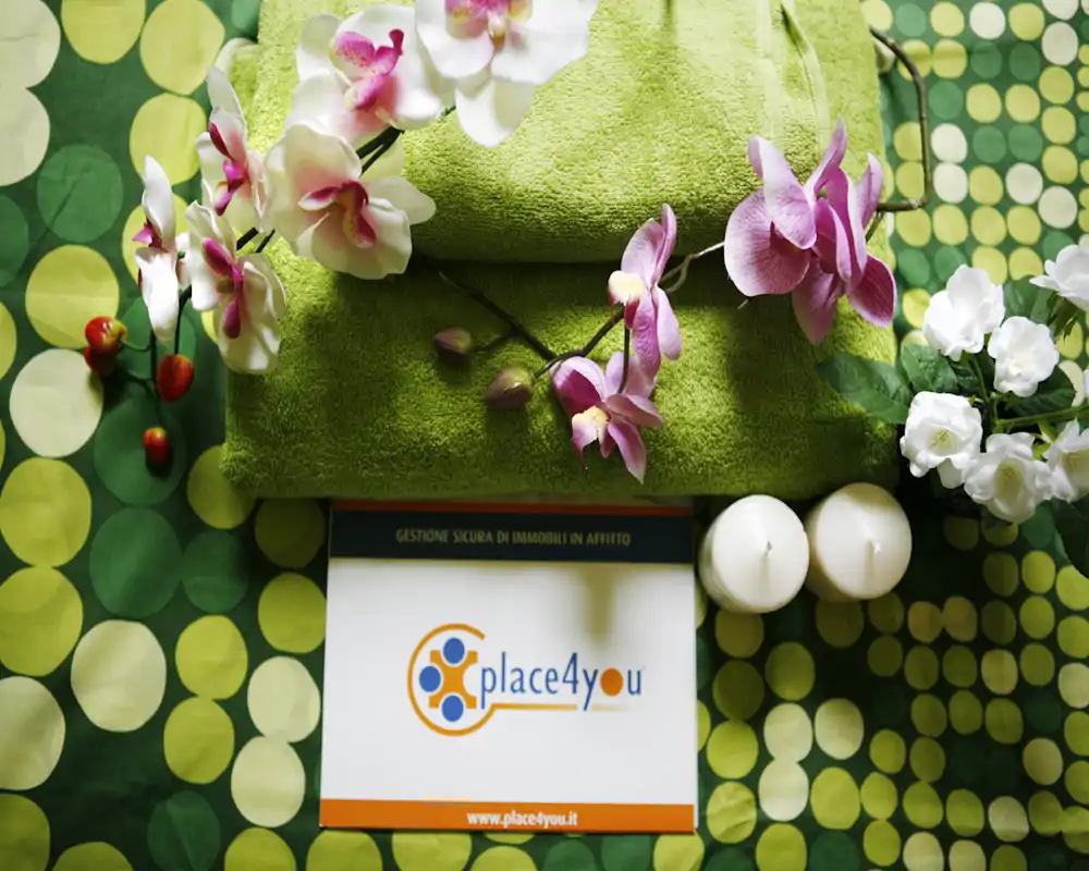 Composición estética en vista cenital con toallas verdes, orquídeas, velas y una tarjeta de marca sobre una tela estampada — probablemente una imagen promocional o un detalle, no una habitación completa.
