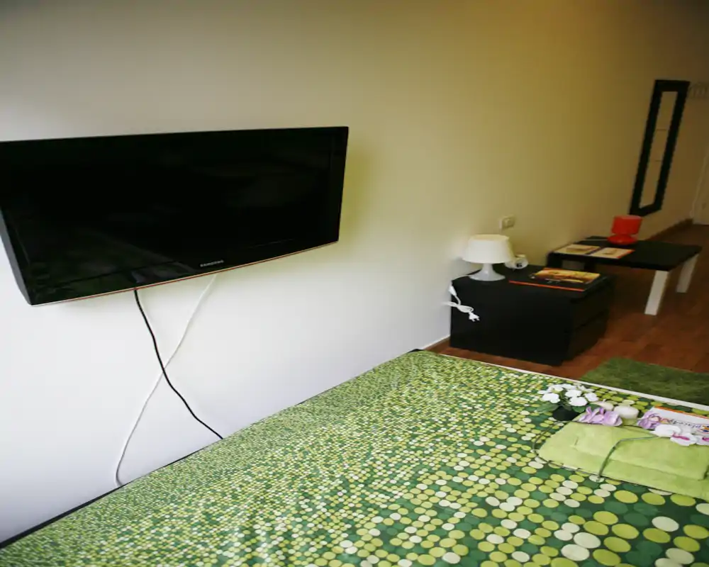 Dormitorio luminoso y ordenado con colcha verde estampada, mesita con lámpara y televisor en la pared — un espacio para dormir acogedor y bien preparado.