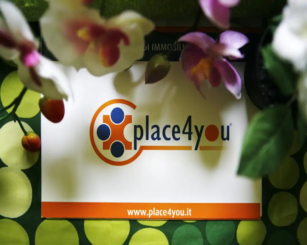 La imagen muestra un primer plano de un cartel impreso con el logotipo 'place4you' y la URL del sitio, parcialmente enmarcado por flores decorativas y un fondo con patrón. No se ven elementos que identifiquen una habitación o función interior.