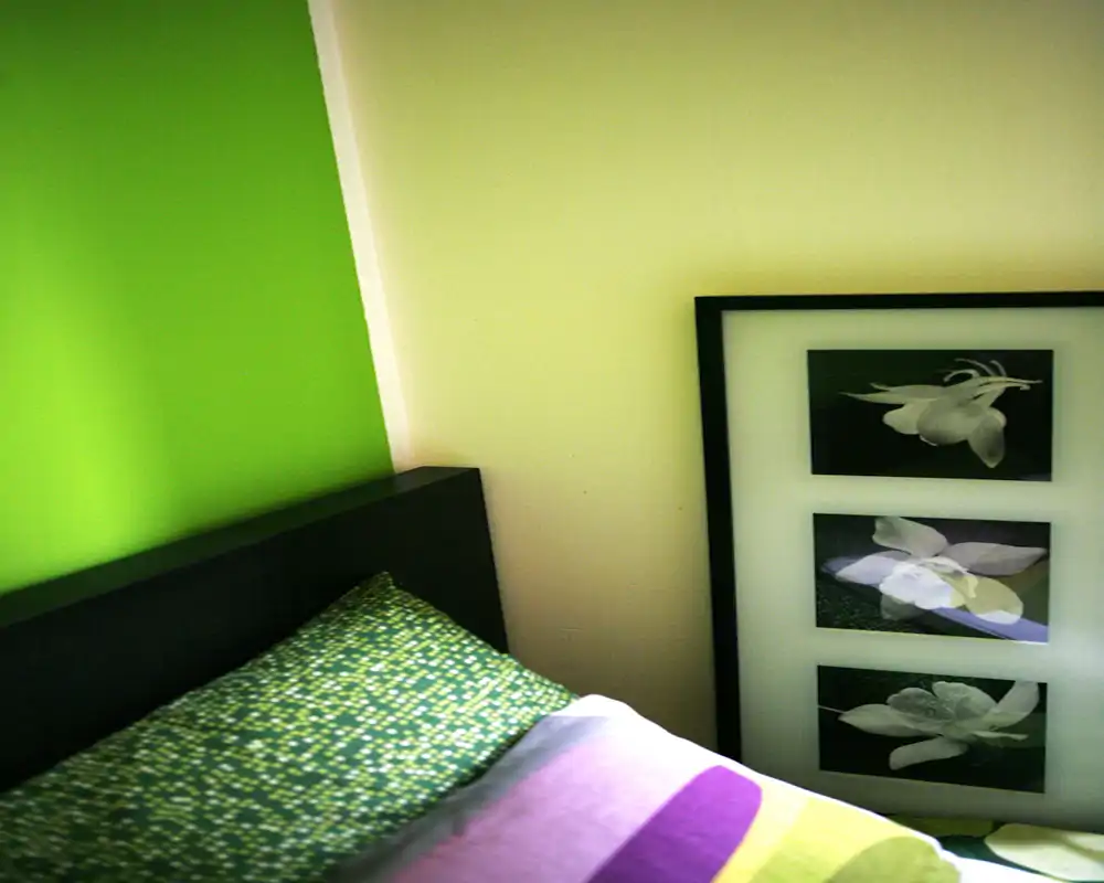 Rincón de dormitorio moderno y luminoso con almohada estampada y edredón colorido sobre una cabecera visible; cuadro contemporáneo apoyado en la pared.