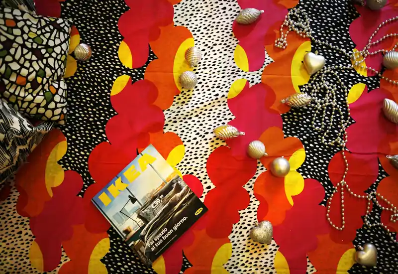 Close-up view of a bedspread with colorful patterned bedding, decorative pillows and small ornaments (IKEA catalog visible). The shot emphasizes the textile and decorative details of a bedroom setting.