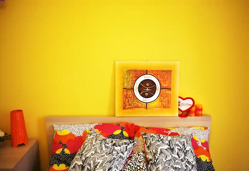 Bright, colorful bedroom with a wooden headboard, patterned pillows and vibrant yellow accent wall with artwork — cozy and well-lit.