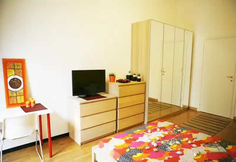 Bright, colorful bedroom with a patterned double bed, wooden dressers and a mirrored wardrobe providing depth. Clean, modern styling with decorative accents and a small desk area.