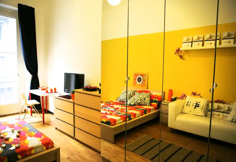 Bright, cozy bedroom with a patterned double bed, mirrored wardrobe, dresser and a small desk by the window. Warm yellow accent wall and colorful textiles create a welcoming atmosphere.