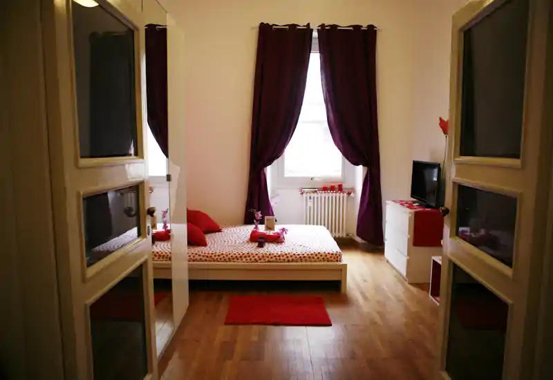 Sunny, stylish bedroom with a low platform bed dressed in patterned linens, rich purple curtains and red accents; wood flooring and natural light create a warm, inviting atmosphere.