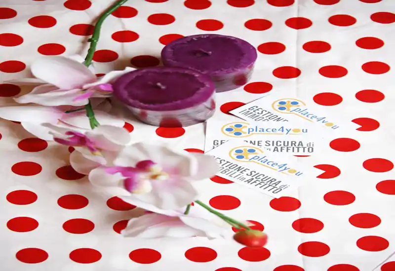 Primer plano de elementos decorativos: dos velas moradas, una rama de orquídea artificial y tarjetas con marca sobre una tela blanca de lunares rojos. La imagen destaca el estilismo y el branding más que una habitación concreta.