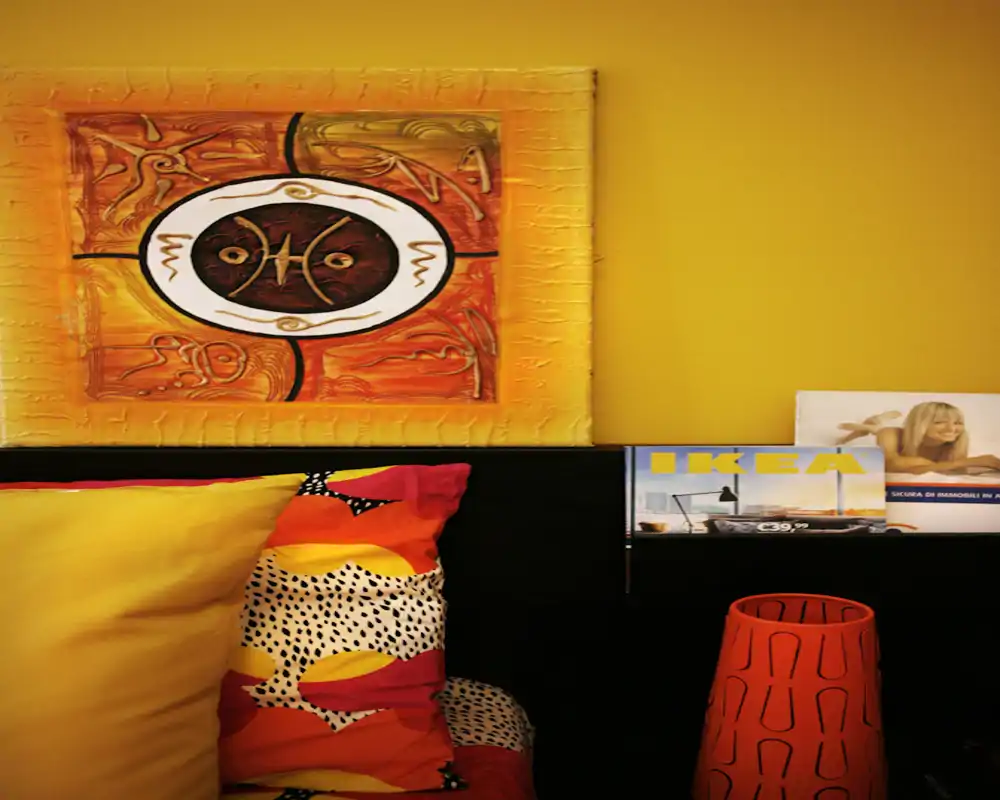 Rincón de dormitorio acogedor con cojines coloridos, paredes amarillas cálidas y un cuadro decorativo sobre la cabecera. La composición resalta los textiles y la iluminación ambiental, perfecta para un anuncio de alquiler.