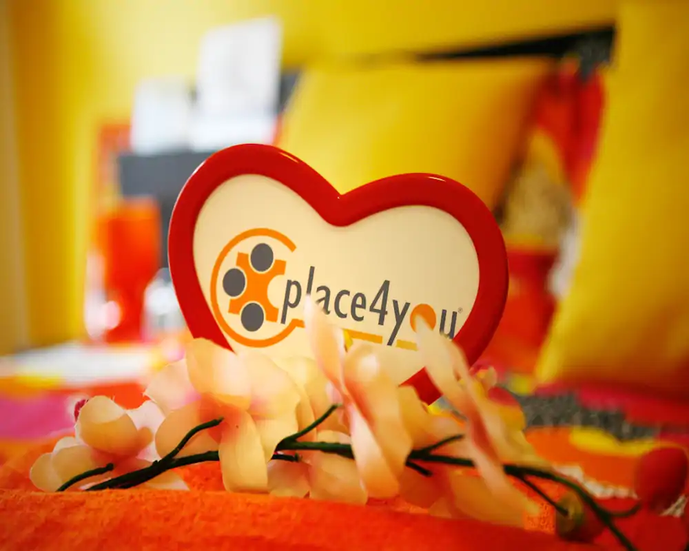 Toma en primer plano decorativa sobre una cama con almohadas amarillas y detalles florales; un letrero en forma de corazón «place4you» enfocado y la cama desenfocada al fondo.