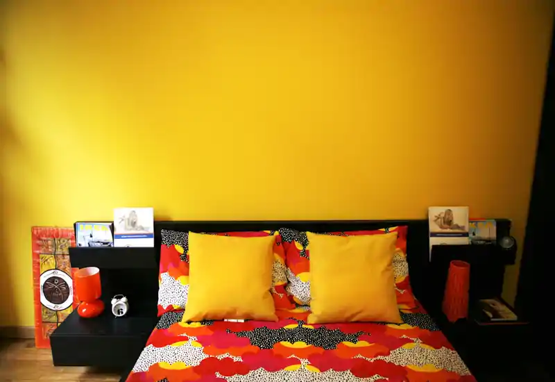 Bright, stylish bedroom with a bold yellow accent wall and colorful patterned bedding. Two matching yellow pillows, a low black headboard with built-in side shelves, and warm wooden flooring create a cheerful, modern atmosphere.