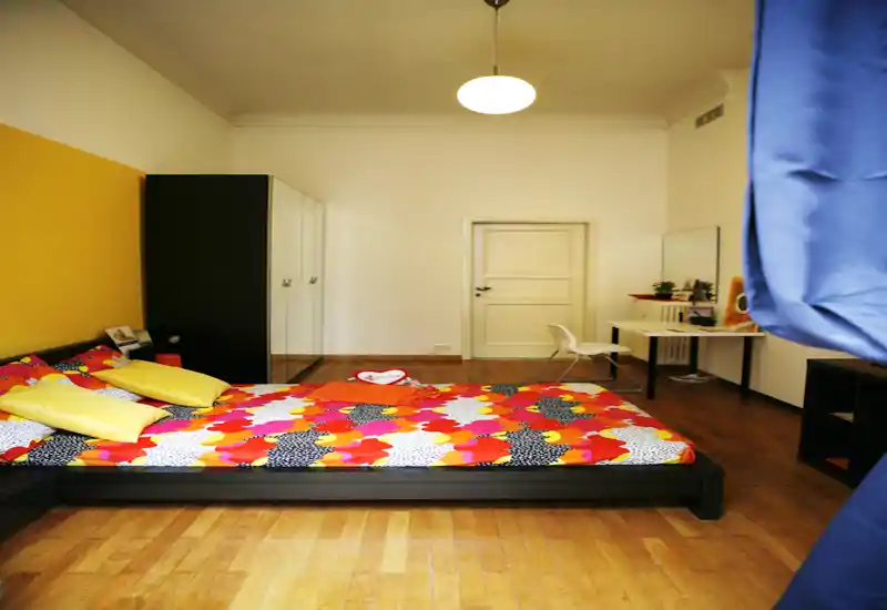 Spacious, well-lit bedroom with a low platform bed featuring colorful bedding, hardwood floors and a simple work desk at the back — bright and inviting for guests.