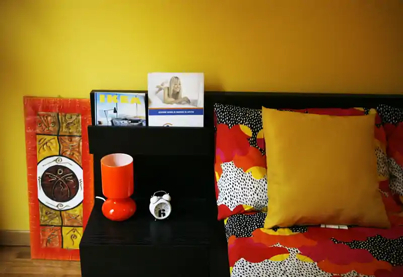 Bright, colorful bedroom corner with patterned bedding, a large yellow cushion and a bedside table styled with a lamp and alarm clock — warm, well-composed shot suitable for listings.