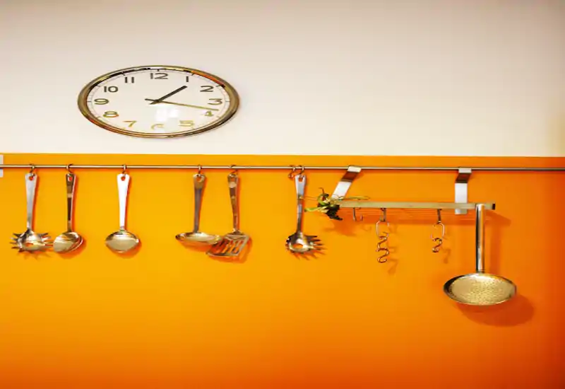 Pared de cocina luminosa y moderna con utensilios de acero colgados en una barra sobre un fondo naranja cálido y un reloj arriba, que crea un aire vivo y con estilo.
