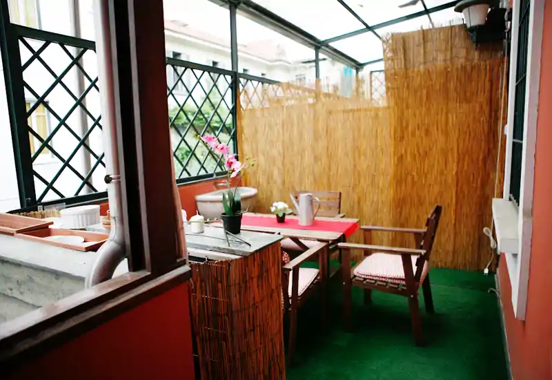 Balcón cubierto con pequeña mesa comedor, dos sillas con cojines, plantas decorativas y pantalla de cañas para privacidad — un rincón exterior acogedor para tomar café o comer.