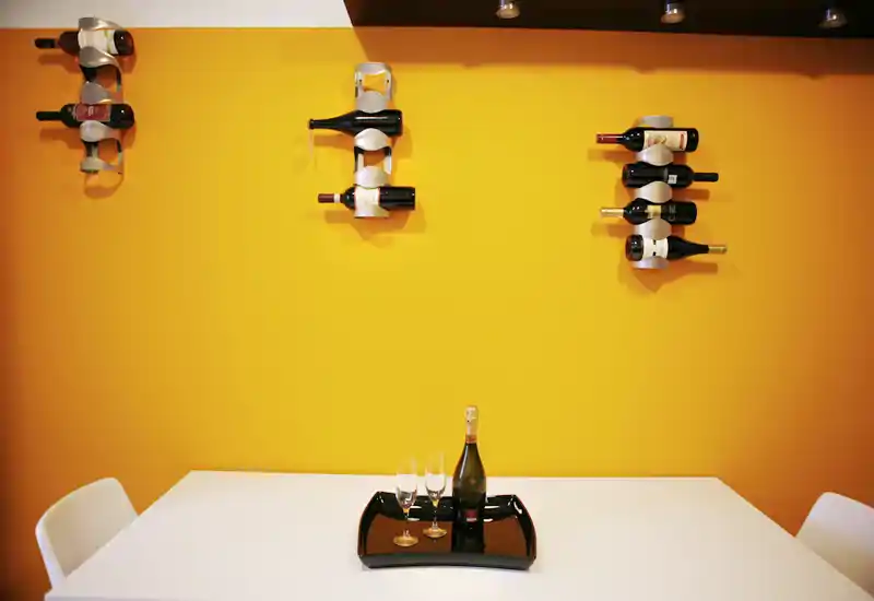 Rincón de comedor contemporáneo con pared amarilla brillante, mesa y sillas blancas minimalistas y botelleros decorativos; una botella y copas sobre una bandeja añaden un toque acogedor.