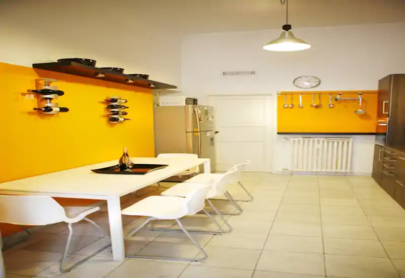 Zona de comedor moderna y luminosa integrada en la cocina: mesa blanca y sillas contemporáneas frente a paredes naranja cálidas, botelleros de pared y utensilios colgados que crean un espacio funcional y con estilo.