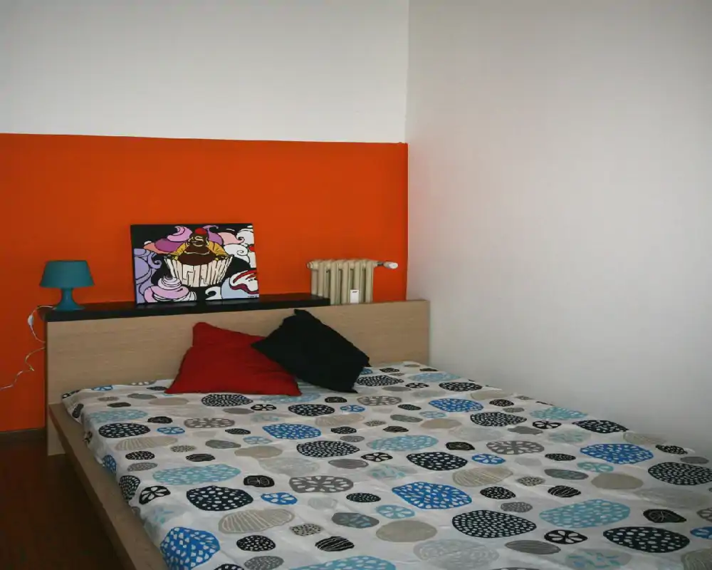 Camera da letto moderna e luminosa con copriletto a motivo, due cuscini decorativi e una parete arancione di accento. Una lampada da comodino e un piccolo quadro sullo schienale creano un’atmosfera contemporanea e vivace.