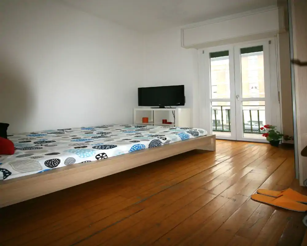 Camera da letto luminosa e semplice con letto su piattaforma in legno, biancheria a fantasia, luce naturale da una finestra alta e una piccola TV su un mobile basso.