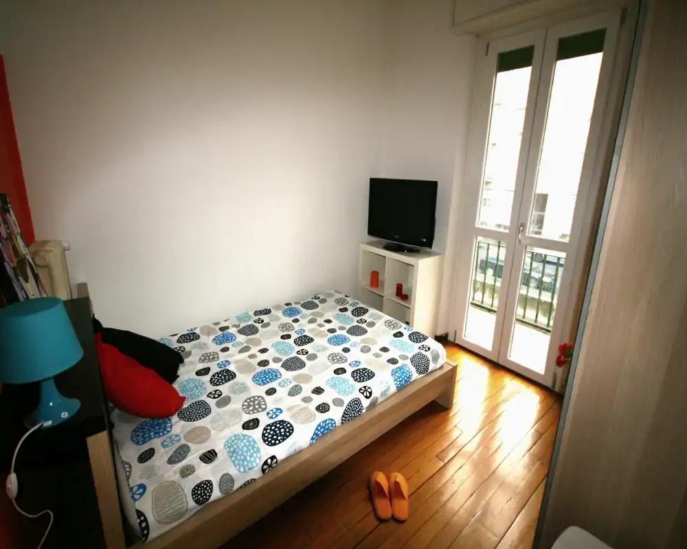 Camera da letto compatta e luminosa con letto singolo, copripiumino a fantasia e porta finestra che illumina l'ambiente. Pavimento in legno, mobile TV e arredo semplice e moderno.