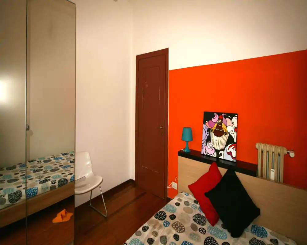 Camera da letto moderna e luminosa con parete arancione, biancheria a motivi e cuscini decorativi; mensola comodino con lampada e quadro colorato, armadio con ante a specchio che riflette parte del letto.