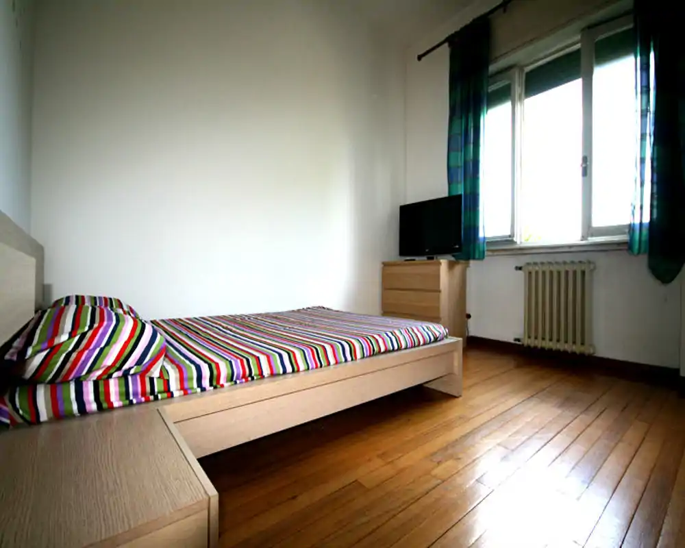 Dormitorio luminoso y minimalista con una cama baja de madera con ropa de cama a rayas coloridas, gran ventana con luz natural y suelos de madera cálida.