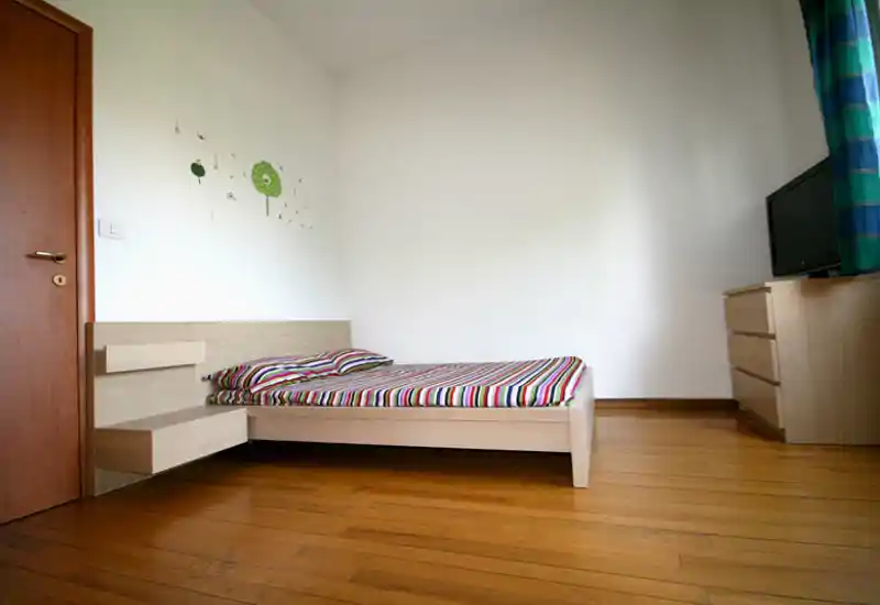 Dormitorio luminoso y minimalista con cama baja de madera y ropa de cama a rayas, suelo de madera y cómoda con televisor — sencillo, limpio y moderno.