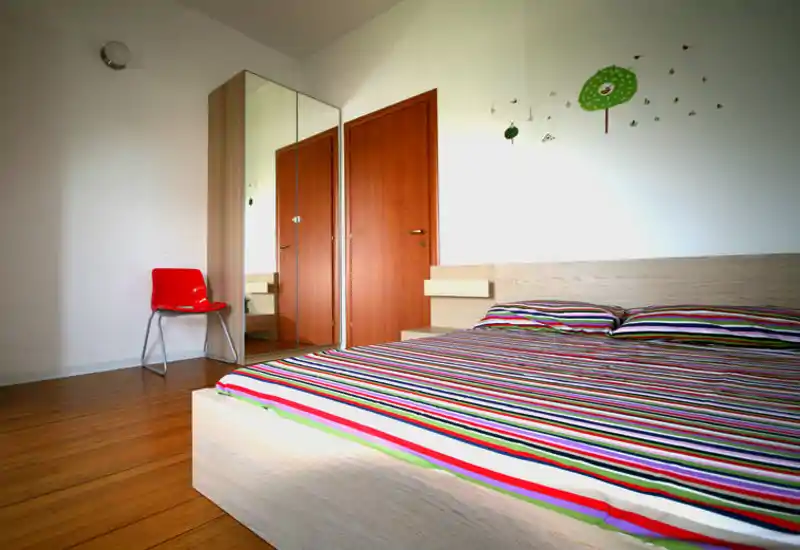 Dormitorio moderno y luminoso con cama baja de madera y ropa de cama a rayas coloridas, armario con espejo y decoración sencilla que aporta sensación de amplitud.