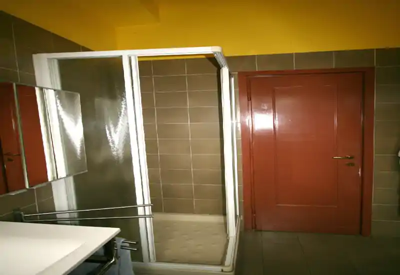 Baño compacto con cabina de ducha en esquina, paredes alicatadas, espejo y lavabo; techo amarillo y puerta rojiza aportan carácter.