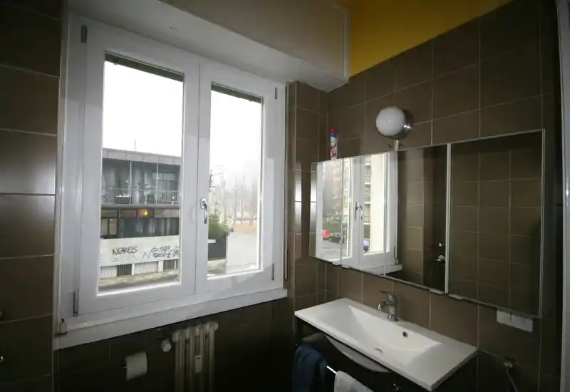 Pequeño baño con lavabo blanco, armario con espejo y una gran ventana que aporta luz natural.