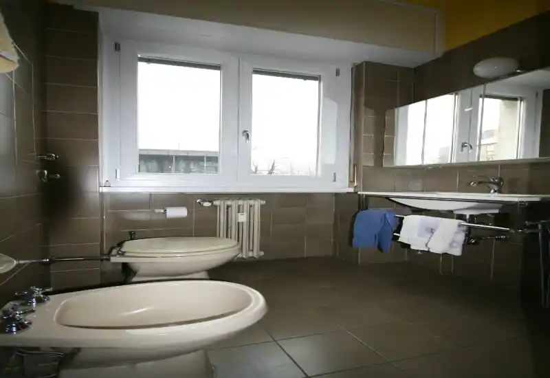 Baño luminoso y ordenado con ventana, inodoro, bidé y lavabo con espejo. Baldosas de tono neutro y luz natural que lo hacen práctico y funcional.