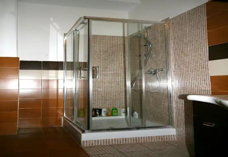 Baño compacto con cabina de ducha de cristal, columna de ducha empotrada y azulejos mosaico; productos de aseo visibles en el plato de ducha.