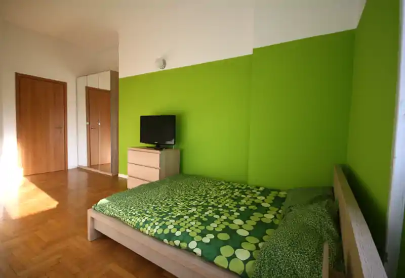 Camera da letto moderna e luminosa con letto matrimoniale in legno e biancheria a fantasia verde, comò con TV e armadio con specchio vicino alla porta. La luce naturale e la parete verde conferiscono un’atmosfera fresca.