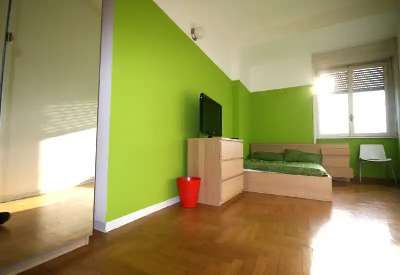 Camera da letto luminosa e moderna con letto basso in legno e comò coordinato su una parete verde brillante; grande finestra che illumina l’ambiente e arredo semplice e minimalista.