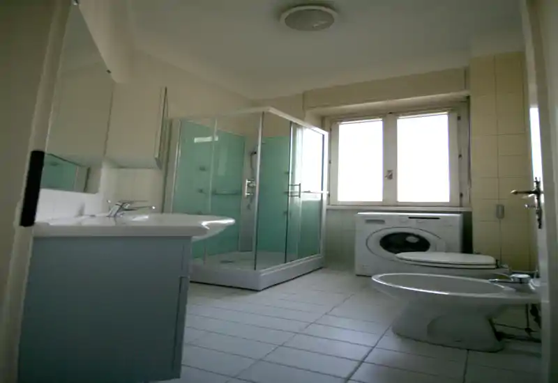 Bagno compatto e luminoso con cabina doccia, lavabo con mobile, bidet, WC e lavatrice sotto la finestra. Piastrelle neutre e luce naturale rendono l'ambiente pulito e funzionale.