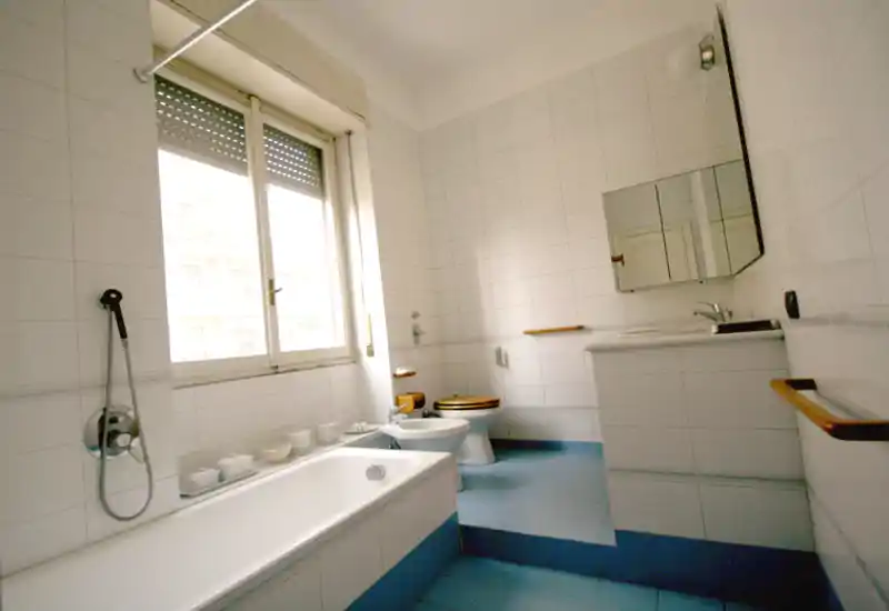 Bagno luminoso e spazioso con vasca, bidet, WC e mobile lavabo. Piastrelle bianche alle pareti, pavimento azzurro e una grande finestra che garantisce molta luce naturale.