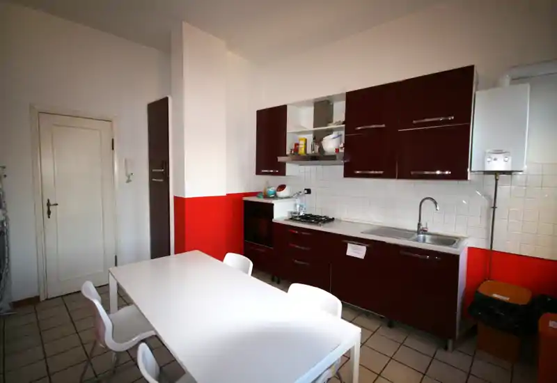 Cucina moderna e luminosa con mobili bordeaux lucidi, piano cottura a gas, forno integrato e tavolo bianco con sedie — pulita, ben illuminata e pronta all'uso.