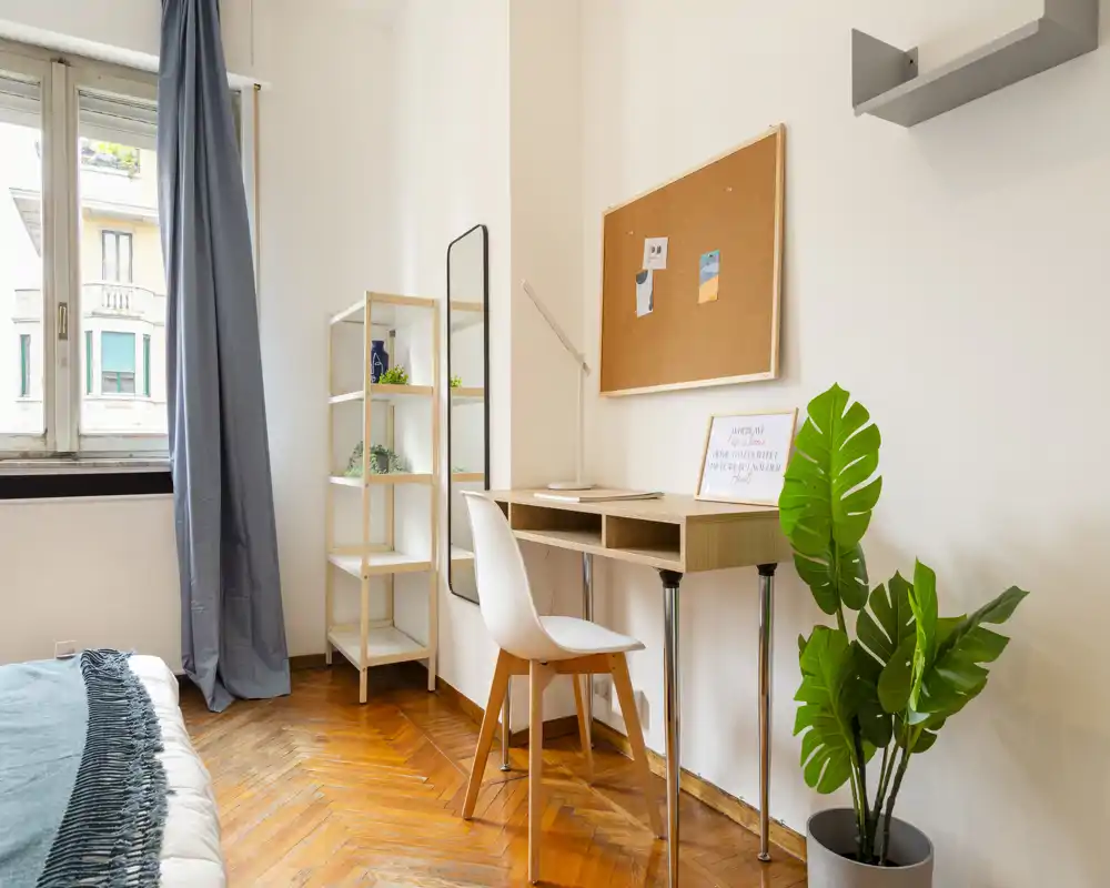 Angolo camera luminoso e curato: letto parzialmente visibile a sinistra, scrivania in legno con sedia, bacheca decorativa e pianta in vaso — perfetto per chi lavora e riposa.