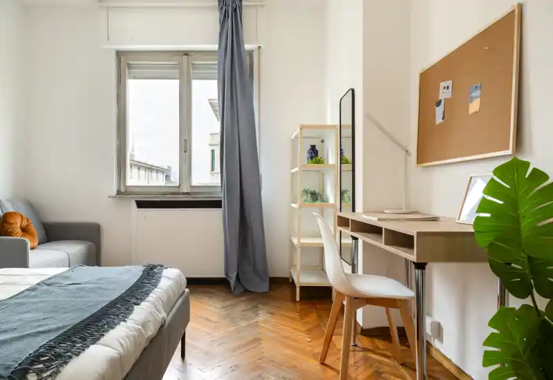Dormitorio luminoso y acogedor con una cama cómoda, un escritorio de trabajo y luz natural de una gran ventana; suelos de madera cálidos y decoración moderna sencilla.