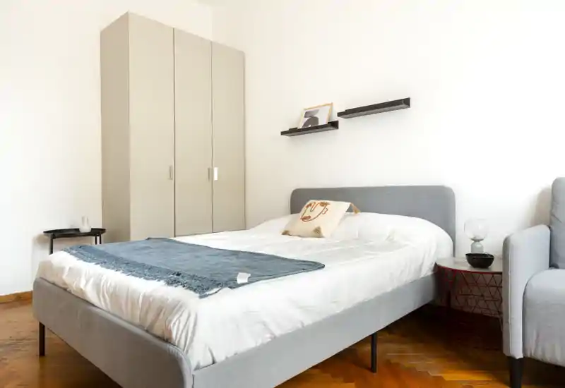 Dormitorio moderno y luminoso con cama tapizada en gris, ropa de cama blanca y manta turquesa; armario y estantes ofrecen almacenamiento y un aspecto acogedor y minimalista.