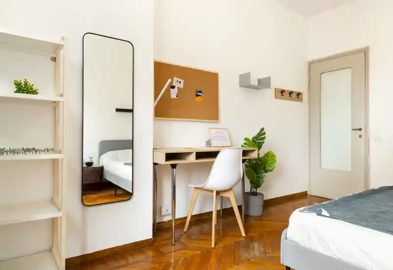 Dormitorio moderno y luminoso con la cama bien hecha, un escritorio y silla junto a la pared, espejo de cuerpo entero y planta en maceta — ideal para estudiante o una sola persona.