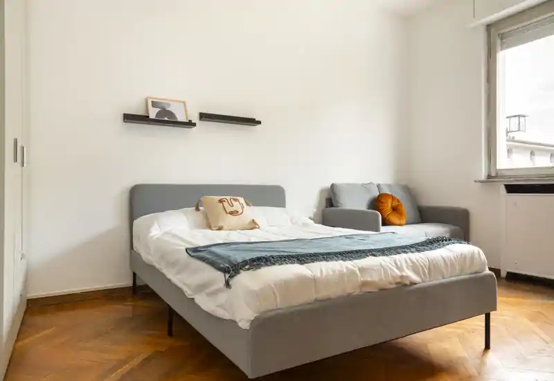 Camera da letto luminosa e moderna con letto matrimoniale con testiera rivestita grigia, biancheria bianca e plaid turchese; poltrona grigia con cuscino arancione vicino alla finestra per un angolo lettura accogliente.